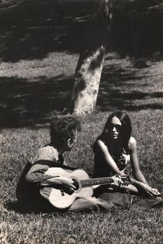 two people sitting in the grass playing guitar and another person standing next to them with a tree behind them