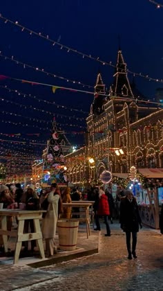 people are walking around an outdoor christmas market at night with lights strung across the street