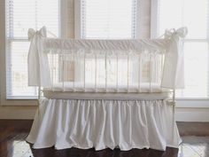 a baby crib with white ruffled bedding in front of two large windows