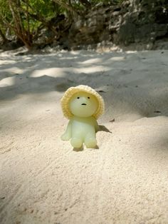 a small toy bear wearing a yellow hat on top of a sandy beach next to trees