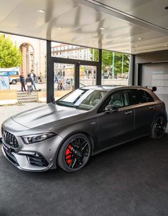 a silver car parked in front of a glass building with people walking around it and onlookers