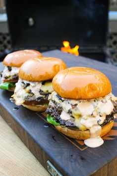 two hamburgers with cheese and other toppings sit on a cutting board next to an open fire