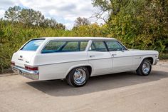 an old white station wagon parked in a parking lot next to some bushes and trees