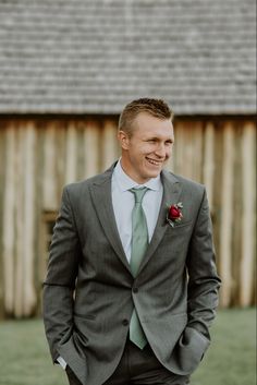 a man in a gray suit and green tie smiles at the camera while standing next to a barn