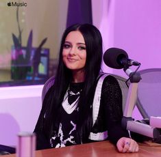 a woman sitting at a desk with a microphone in front of her and an apple computer behind her