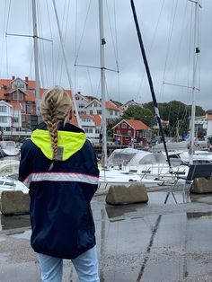 a woman standing in the rain next to sailboats at a marina with houses and buildings behind her