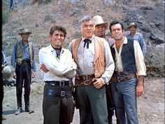 four men standing together in front of a rocky area with one man wearing a vest and tie