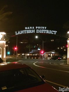 a red car driving down a street next to a tall white building with a sign that reads dana point lanternern district