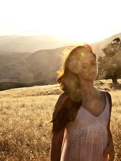 a woman standing on top of a grass covered field next to a tree and mountains