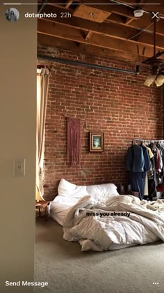 an unmade bed sitting in front of a brick wall with clothes hanging on it