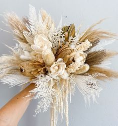 a hand holding a bouquet of dried flowers and grasses in front of a white wall