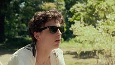 a young man wearing sunglasses sitting on a bench in front of some trees and bushes