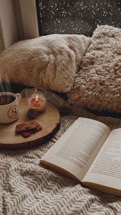 an open book sitting on top of a bed next to a cup of coffee and candle