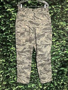 a pair of camouflage pants hanging on a green bushy wall in front of a white hanger