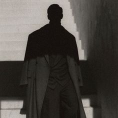 a black and white photo of a man in a trench coat walking up some stairs