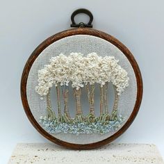 an embroidery project with trees and bushes in the center on a wooden frame, sitting on top of a white surface