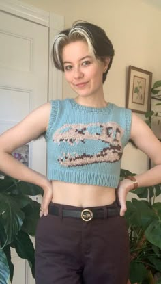 a woman standing in front of a potted plant wearing a blue sweater and black pants