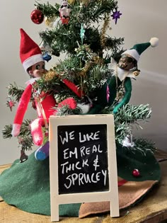 an elf is sitting in front of a christmas tree with a sign that says we like em real trick 4 sprucey