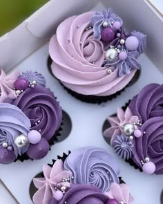 four cupcakes with purple frosting and flowers on them in a white box