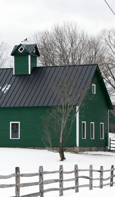 a green barn with a black roof and white trim in the winter snow near a wooden fence