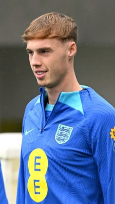 a young man wearing a blue shirt with yellow and white numbers on the chest, standing in front of a soccer field