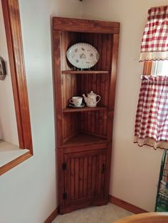 a wooden corner cabinet in the corner of a room with a checkered window curtain