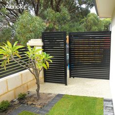 a black gate is open in front of a small tree and shrubbery on the side of a house