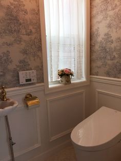 a white toilet sitting under a window next to a sink in a room with floral wallpaper