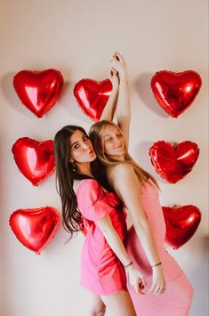 two young women are posing in front of red heart - shaped balloons on the wall