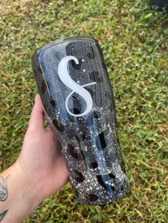 a hand holding up a wine glass with the letter s on it in front of some grass