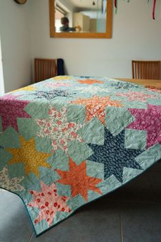 a bed with a colorful quilt on top of it next to two wooden chairs and a mirror