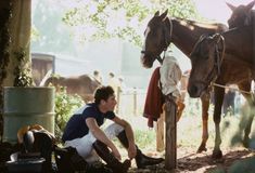 a man sitting on the ground next to a horse and another person standing near by