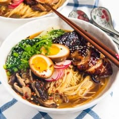 two bowls of ramen with chopsticks and an egg on the side, sitting on a blue checkered tablecloth