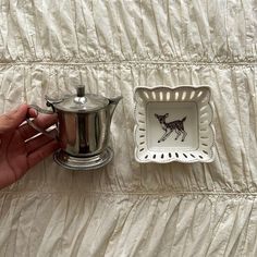 a person holding a silver tea pot next to a white plate with a deer design on it