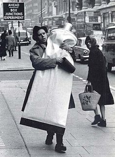 an old black and white photo of a woman carrying bags down the street with other people walking by