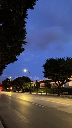 an empty street at night with the lights on and trees lining the road in front of it