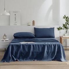 a bed with blue sheets and pillows in a white room next to a potted plant