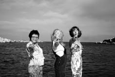 three women standing next to each other in front of the ocean pointing at something with their fingers