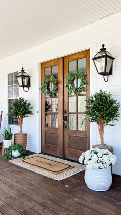 two potted plants sit on the front porch of a white house with wooden doors