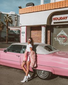 a woman standing next to a pink car in front of a building with palm trees