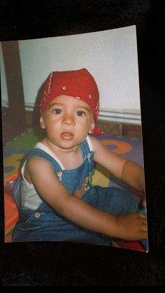 an old photo of a baby wearing a red hat and blue overalls sitting on a bed