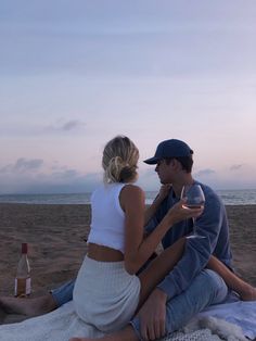 a man and woman sitting on the beach next to each other with wine in their hands