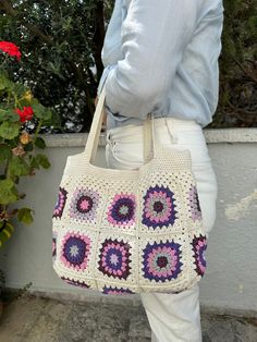 a woman carrying a crocheted bag in front of a flower pot with red flowers