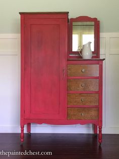 a red armoire sitting next to a white vase on top of a wooden table