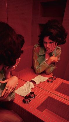 two women sitting at a red table with papers on it and one is touching her face