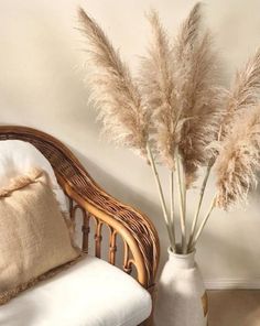 a white vase filled with dry grass next to a wicker chair and throw pillow