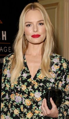 a woman with long blonde hair wearing a black floral shirt and holding a clutch bag