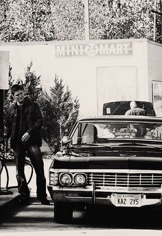 a black and white photo of a man standing next to a car at a gas station