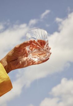 a person holding up a clear glass in front of a blue sky with white clouds