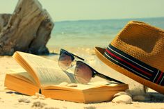 an open book with sunglasses and hat on the beach